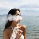 woman wearing white sleeveless top smoking tobacco while standing near blue sea under white and blue skies during daytime