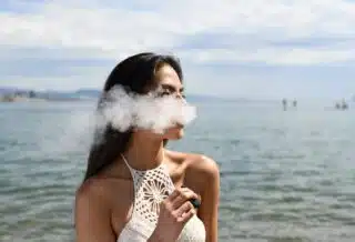 woman wearing white sleeveless top smoking tobacco while standing near blue sea under white and blue skies during daytime