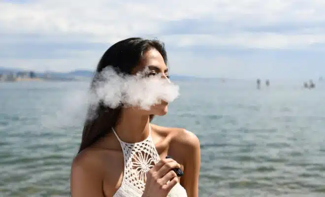 woman wearing white sleeveless top smoking tobacco while standing near blue sea under white and blue skies during daytime